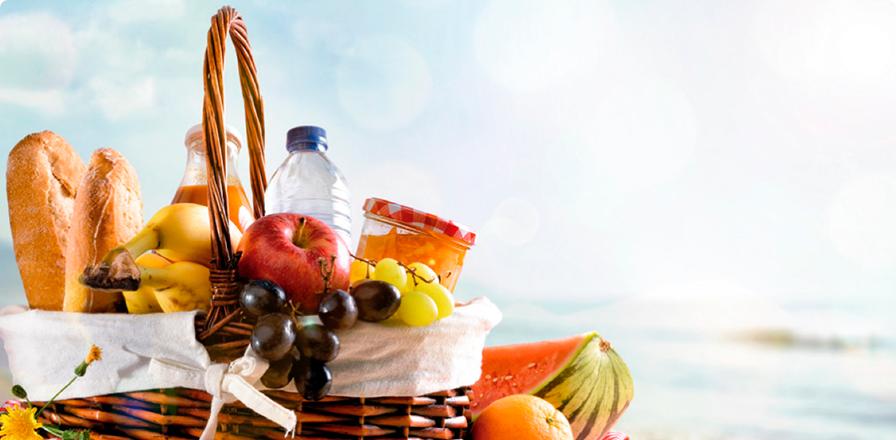 Una cesta de mimbre tipo picnic con alimentos como: pan, fruta, mermelada, zumos y agua