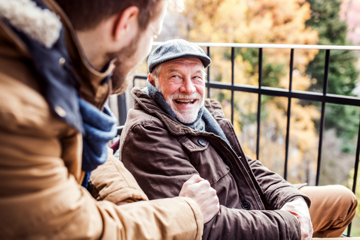 Un hombre mayor sonríe a un hombre joven mientras ambos están sentados en una terraza de un edificio